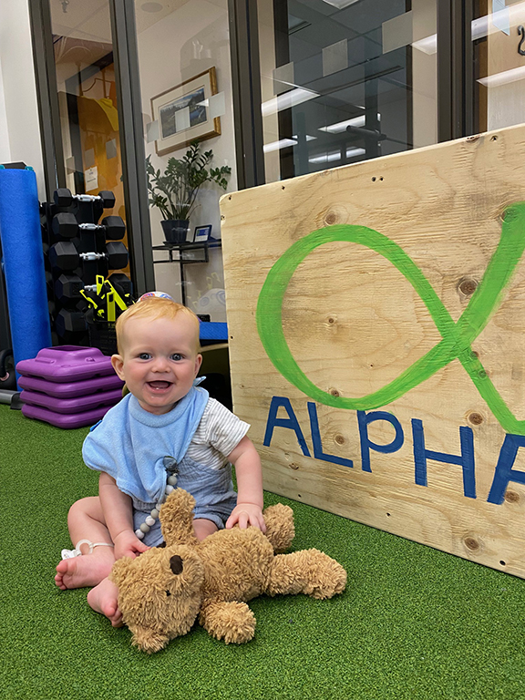 image of baby and teddybear at East clinic in front of wooden board with Alpha logo on it